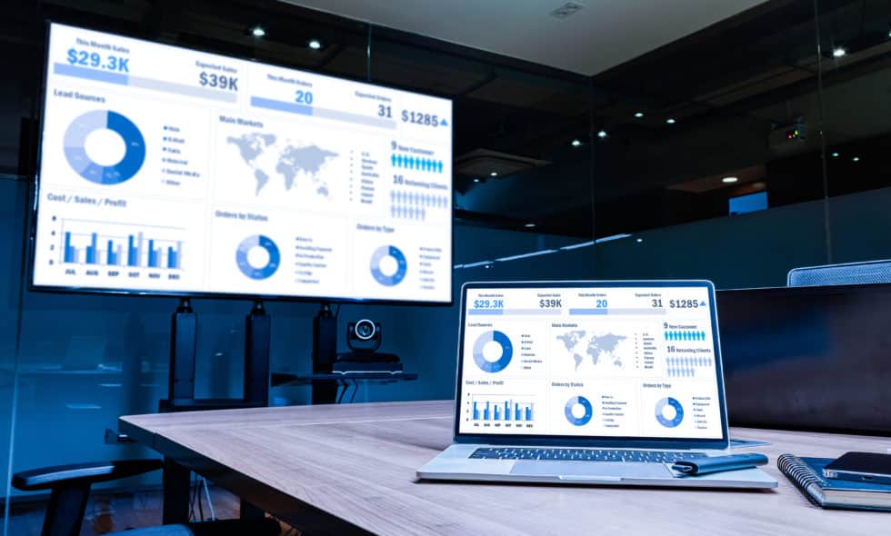 Mock up sales summary slide show presentation on display television and laptop with notebook on table in meeting room